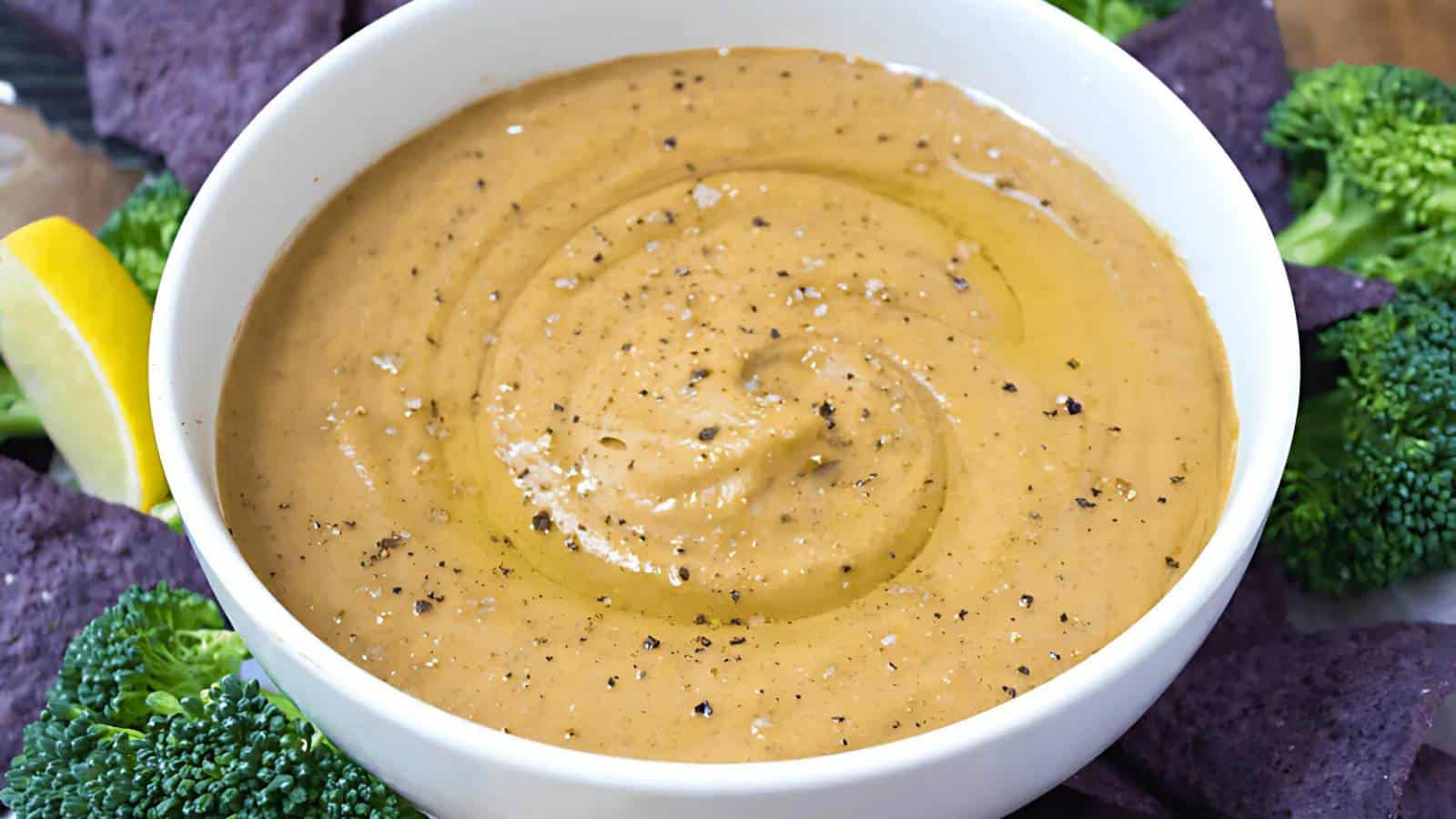 A white bowl filled with a creamy light brown dip, topped with black pepper and olive oil. Surrounded by broccoli and blue corn chips.