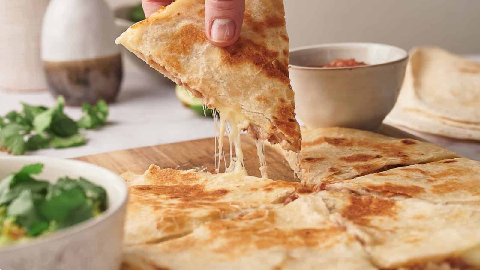 A hand lifts a triangular slice of a cheese quesadilla from a wooden board. Additional bowls with cilantro and salsa are in the background.
