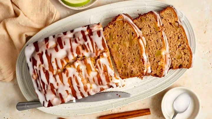Sliced loaf of glazed cinnamon apple bread on an oval plate with a knife, small bowl, green apple slice, and cinnamon sticks nearby.