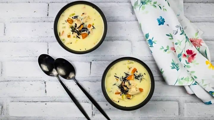 Two bowls of creamy soup with vegetables and herbs, placed on a white brick surface. Two black spoons and a floral napkin are beside the bowls.