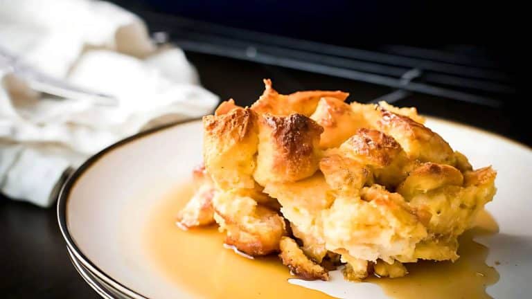A slice of bread pudding topped with syrup sits on a white plate with a dark rim, placed on a dark surface next to a white cloth napkin.