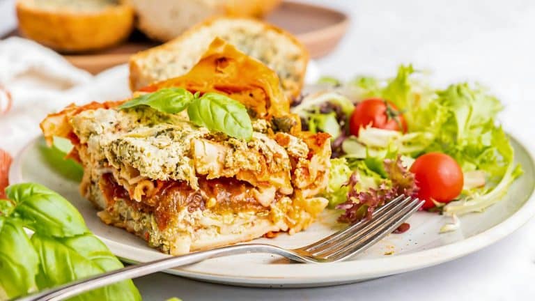 A plate with a slice of spinach lasagna topped with basil, accompanied by a side salad with cherry tomatoes and a slice of bread.