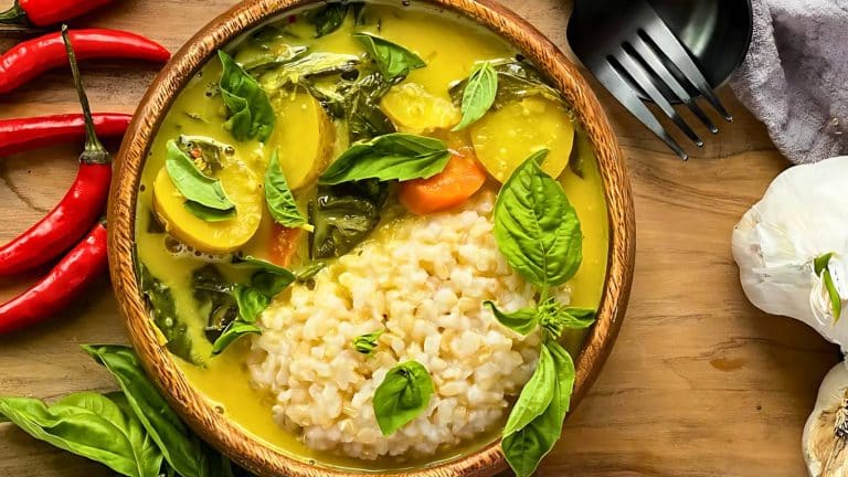 A wooden bowl of curry with rice, assorted vegetables, and fresh basil leaves. Red chili peppers, garlic, and a fork and spoon are placed beside it on a wooden surface.