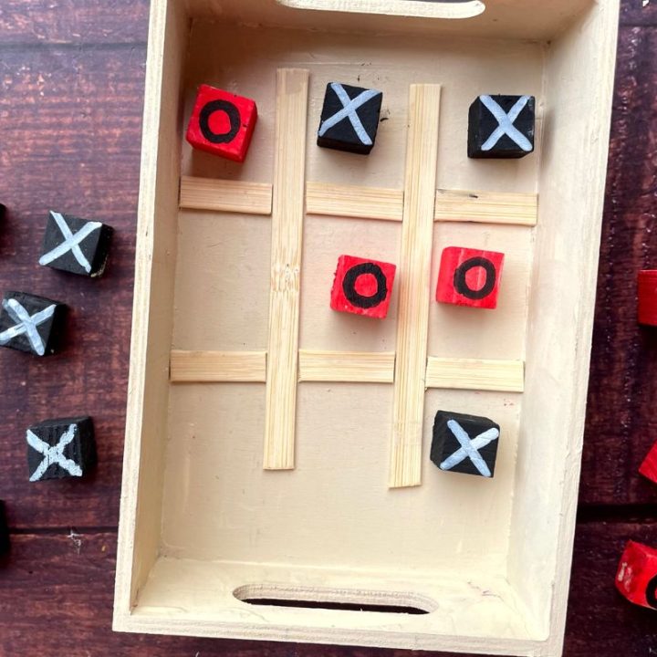 A wooden tic-tac-toe game with black and red blocks, displaying X's and O's. The game board is made of a small wooden tray with dividers. Some blocks are placed inside, while others are scattered outside.