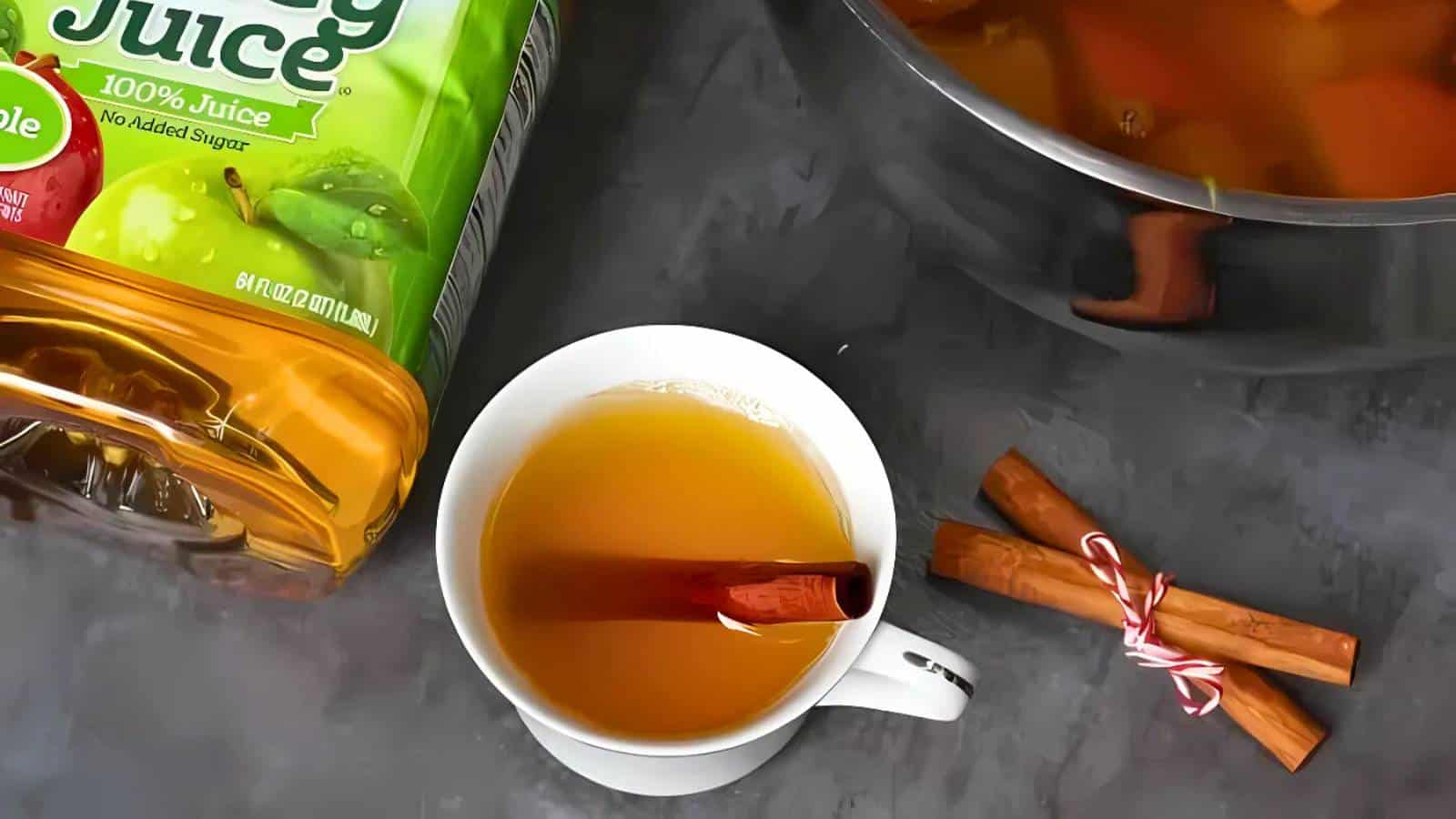 A cup of apple juice with a cinnamon stick on a table, next to a bottle of apple juice and two cinnamon sticks tied together.