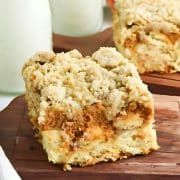 Two slices of crumbly apple coffee cake on wooden boards, with cinnamon sticks and milk bottles in the background.