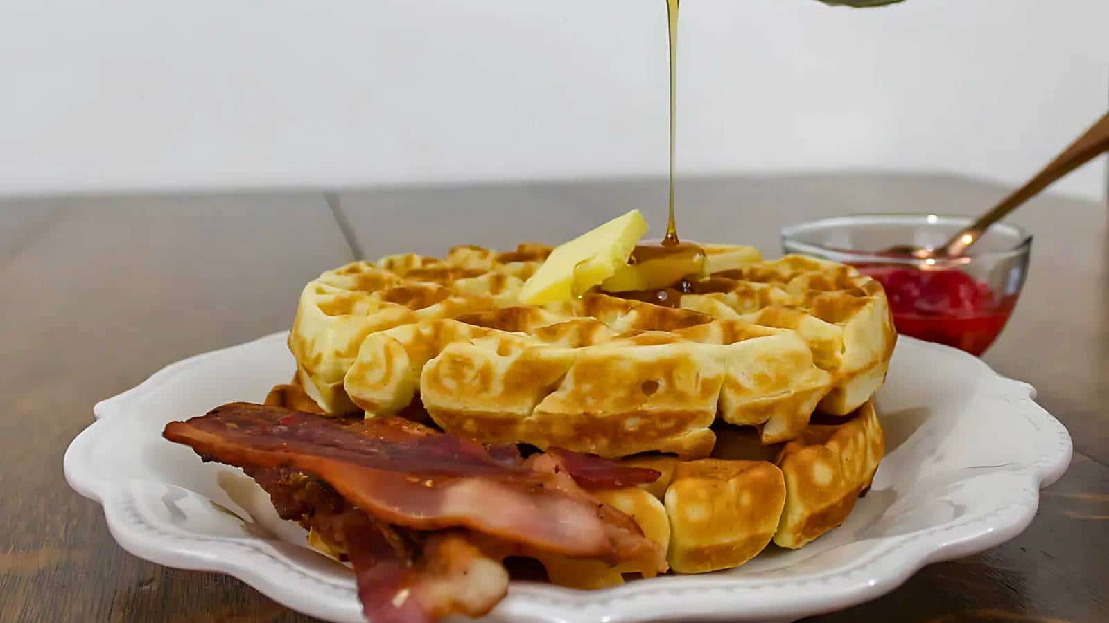 A stack of waffles topped with butter and syrup on a white plate, accompanied by bacon. A small bowl of red jam is in the background.