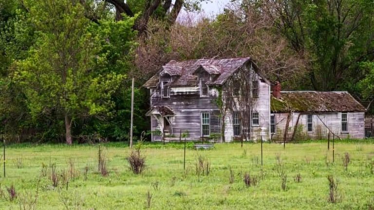 An old, weathered wooden house with a sagging roof stands in a grassy field surrounded by tall trees.