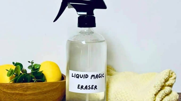 A spray bottle with a label "Liquid Magic Eraser" is placed next to a wooden bowl containing lemons and greenery, and a folded beige towel.