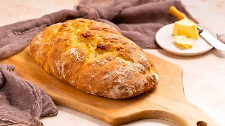 A loaf of crusty bread on a wooden board, accompanied by a small plate with butter and a butter knife, set against a textured, soft, gray fabric.