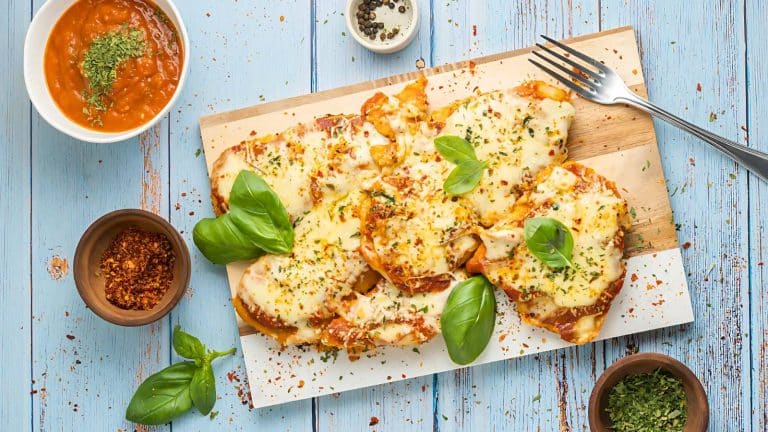 Cheesy garlic bread topped with herbs and basil on a wooden board, surrounded by bowls of sauce, pepper, and chili flakes on a blue wooden table. A fork lies nearby.