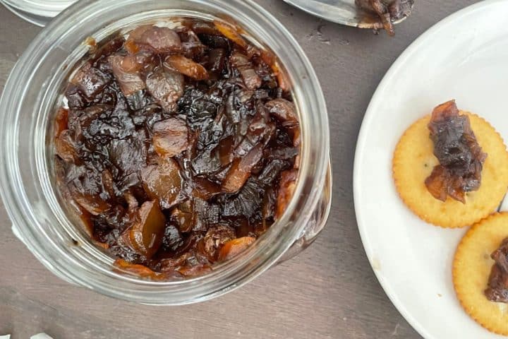 A jar of dark balsamic onion relish sits next to a plate with crackers generously topped with the same rich jam, all elegantly placed on a rustic wooden surface.