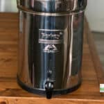 A stainless steel Berkey water filter dispenser, perfect for eco friendly gifts, sits on a wooden table with a faucet at the bottom and a logo on the front.