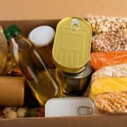 A cardboard box containing best foods for prepping such as bottles, cans, rice, lentils, chickpeas, oats, cornmeal, and jars.