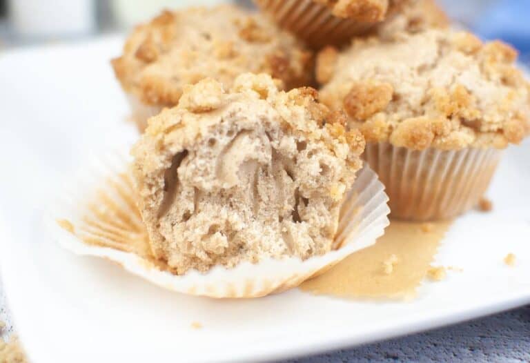 A halved sourdough discard muffin with crumbled topping graces a white plate, accompanied by three whole muffins in the background. The muffin reveals a delightfully crumbly interior.