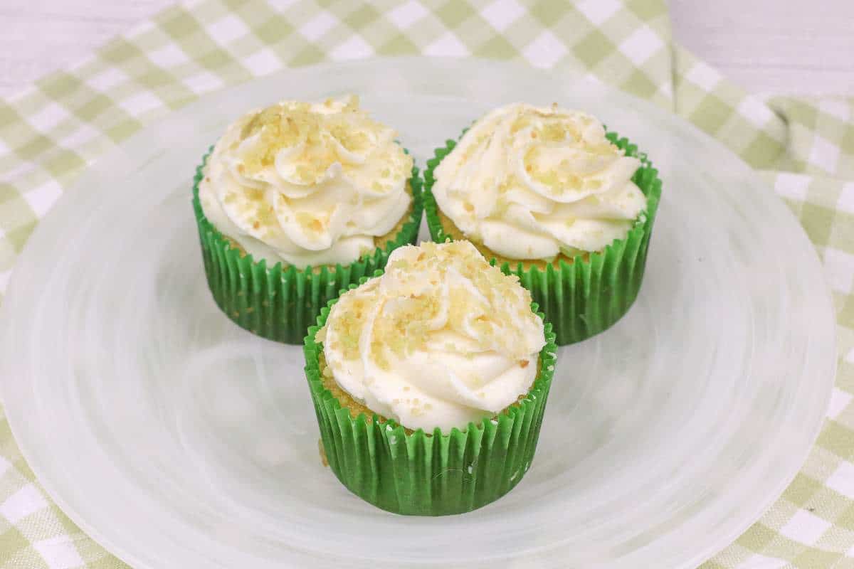 Three coconut cupcakes with lime frosting and sprinkles on a white plate with a green and white checked napkin.