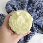 A hand cradles a jar filled with creamy, pale yellow DIY hair butter, set against a backdrop of blue patterned cloth.