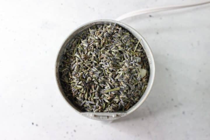 Top view of a grinder filled with dried lavender buds, placed on a speckled white surface.
