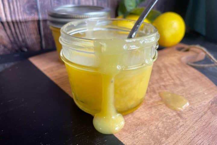 A small glass jar filled with yellow lemon curd, with some of it dripping down the side. A metal spoon is placed inside the jar. Lemons and a covered jar are in the background.