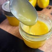 A spoon drips yellow lemon curd into a glass jar on a wooden cutting board with another jar and lemons in the background.