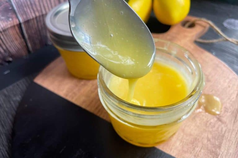A spoon drips yellow lemon curd into a glass jar on a wooden cutting board with another jar and lemons in the background.