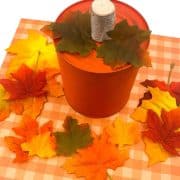 Orange can decorated to resemble a pumpkin, topped with a cork and surrounded by autumn leaves on a checkered surface.