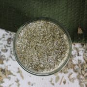 Glass jar filled with eucalyptus bath salts on a white surface with scattered herbs and a green cloth in the background.