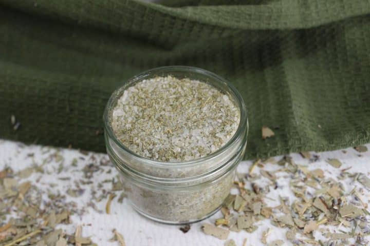 Glass jar filled with a eucalyptus, lavender and peppermint bath salts mixture, placed on a textured green cloth with scattered herbs around.