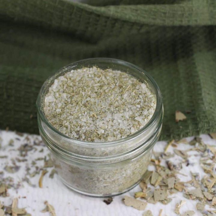 Glass jar filled with a eucalyptus, lavender and peppermint bath salts mixture, placed on a textured green cloth with scattered herbs around.