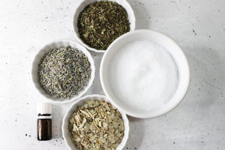 Four bowls containing dried herbs and salt, alongside a small amber bottle, arranged on a white speckled surface.