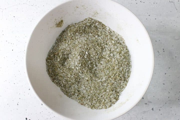 A white bowl filled with a mix of coarse salt and dried herbs on a speckled gray surface.