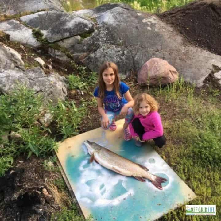 Two young girls outdoors next to a wooden plank with one large fish.