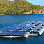 Floating solar panels on a calm body of water, surrounded by tree-covered hills under a clear blue sky.