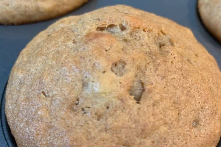 Close-up of a baked muffin in a muffin tin, with a golden-brown top and small cracks visible.