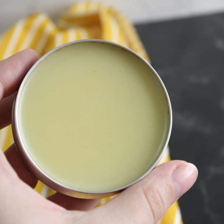 A hand holds an open tin containing a creamy, yellowish balm.