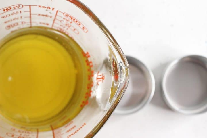 A glass measuring cup filled with yellow liquid is seen from above, next to two silver lids on a white surface.