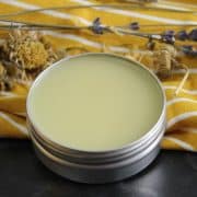 A tin of balm with a smooth surface sits on a yellow striped cloth, surrounded by dried lavender and other herbs.
