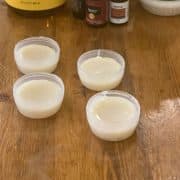 Four small plastic containers filled with orange spiced beeswax lip balm are placed on a wooden table, with several bottles of ingredients seen in the background.