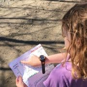 A young girl with wavy brown hair and a watch on her wrist looks at a map and points to a location while standing on a dirt path.
