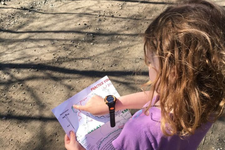 A young girl with wavy brown hair and a watch on her wrist looks at a map and points to a location while standing on a dirt path.