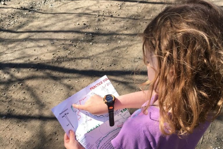 A young girl with wavy brown hair and a watch on her wrist looks at a map and points to a location while standing on a dirt path.