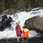 Two young girls looking at rapids by the side of large rocks in the springtime with knapsacks on their backs