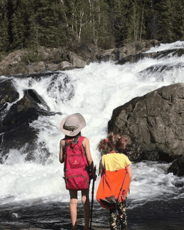 Two young girls looking at rapids by the side of large rocks in the springtime with knapsacks on their backs