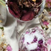 Two glasses sit on a white cloth, one filled with dried flower petals and the other containing salt and small flower petals. Scattered flower petals surround the glasses.