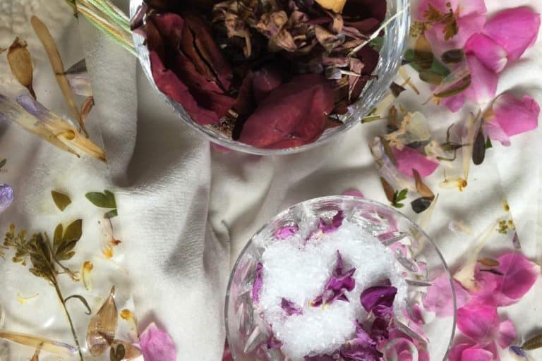 Two glasses sit on a white cloth, one filled with dried flower petals and the other containing salt and small flower petals. Scattered flower petals surround the glasses.