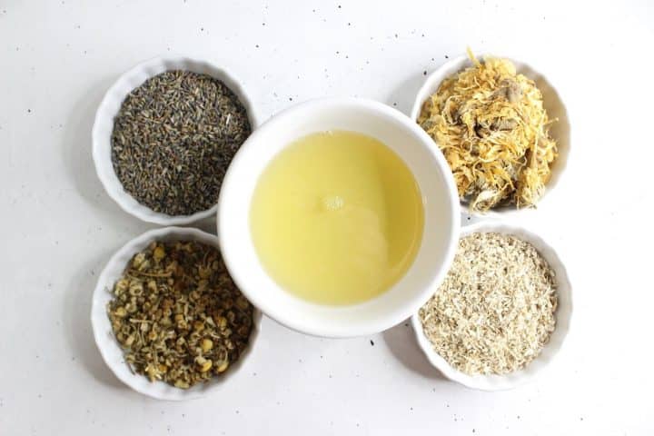 Four small bowls of dried herbs surround a central bowl of yellow liquid on a white surface.