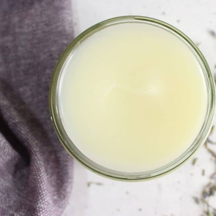 Top view of a glass filled with a light yellow liquid on a white surface, next to a purple fabric and scattered dried lavender flowers.