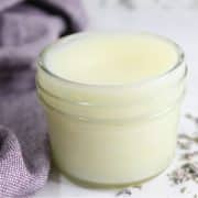 A small glass jar filled with lavender tallow balm sits on a surface with scattered lavender buds and a purple cloth nearby.