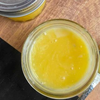 Top view of an opened jar filled with a yellow, creamy substance next to a spoon containing the same substance, on a wooden surface. Another similar jar is partially visible in the background.
