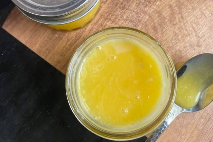 Top view of an opened jar filled with a yellow, creamy substance next to a spoon containing the same substance, on a wooden surface. Another similar jar is partially visible in the background.